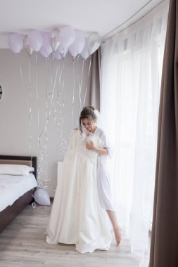 İpek bornozlu genç kadın gelinliğine dokunuyor ve pencerenin yanında dururken gülümsüyor. Düğün günü. Güzellik, moda. En mutlu düğün günü. Her anın tadını çıkar.