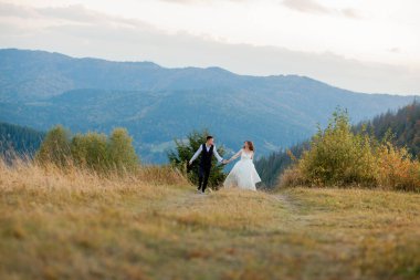 Mutlu muhteşem gelin ve şık damat koşuyor ve eğleniyor, düğün çifti, muhteşem manzaralı dağlarda lüks bir tören.