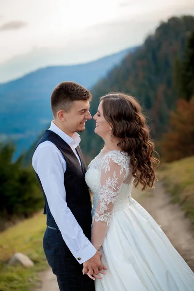 Bella coppia di sposi, sposa e sposo, innamorati sullo sfondo delle montagne. Lo sposo in un bel vestito e la sposa in un abito bianco di lusso. Matrimonio coppia è a piedi — Foto Stock