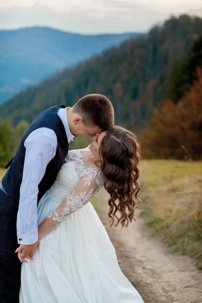 Bella coppia di sposi, sposa e sposo, innamorati sullo sfondo delle montagne. Lo sposo in un bel vestito e la sposa in un abito bianco di lusso. Matrimonio coppia è a piedi — Foto Stock