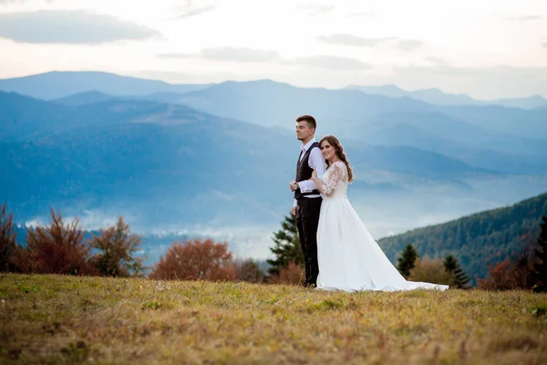 Bella coppia di sposi, sposa e sposo, innamorati sullo sfondo delle montagne. Lo sposo in un bel vestito e la sposa in un abito bianco di lusso. Matrimonio coppia è a piedi — Foto Stock