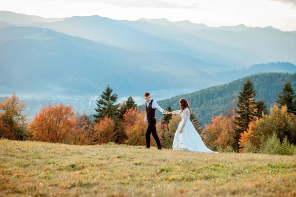 Bella coppia di sposi, sposa e sposo, innamorati sullo sfondo delle montagne. Lo sposo in un bel vestito e la sposa in un abito bianco di lusso. Matrimonio coppia è a piedi — Foto Stock