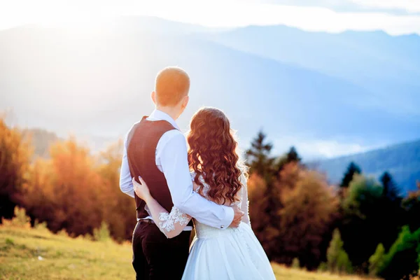 Bella coppia di sposi, sposa e sposo, innamorati sullo sfondo delle montagne. Lo sposo in un bel vestito e la sposa in un abito bianco di lusso. Matrimonio coppia è a piedi — Foto Stock