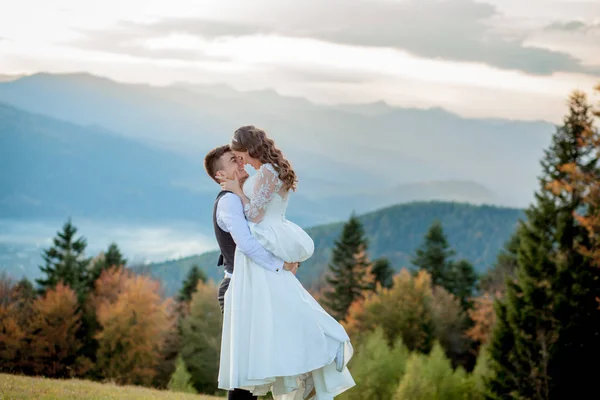 Bella coppia di sposi, sposa e sposo, innamorati sullo sfondo delle montagne. Lo sposo in un bel vestito e la sposa in un abito bianco di lusso. Matrimonio coppia è a piedi — Foto Stock