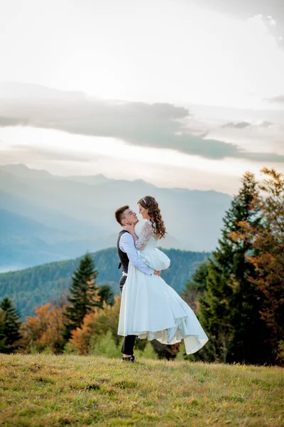 Gyönyörű házaspár, menyasszony és vőlegény, szerelmesek a hegyek hátterében. A vőlegény egy gyönyörű öltönyben és a menyasszony egy fehér luxus ruhában. Az esküvői pár sétál. — Stock Fotó