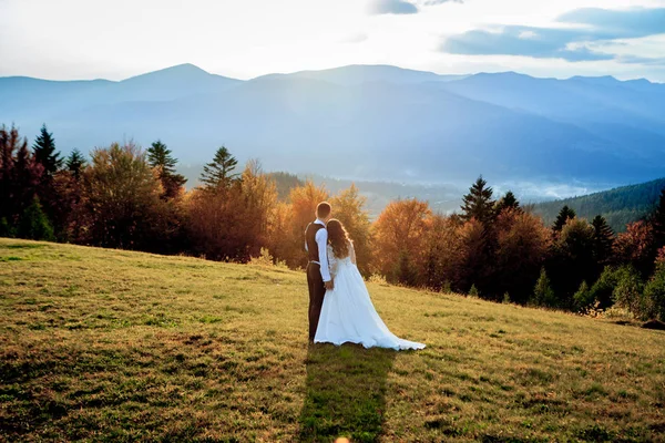 Bella coppia di sposi, sposa e sposo, innamorati sullo sfondo delle montagne. Lo sposo in un bel vestito e la sposa in un abito bianco di lusso. Matrimonio coppia è a piedi — Foto Stock