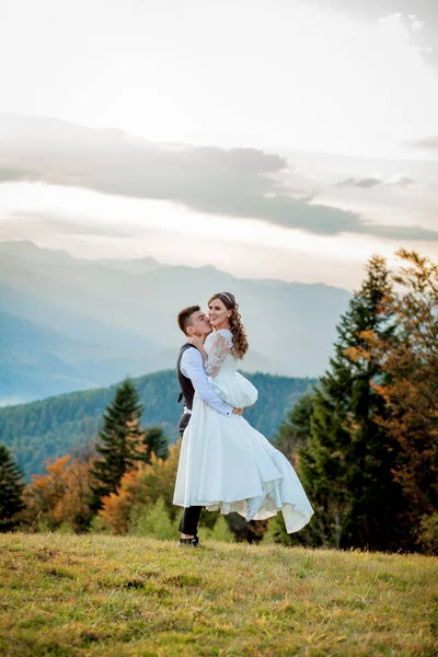 Gyönyörű házaspár, menyasszony és vőlegény, szerelmesek a hegyek hátterében. A vőlegény egy gyönyörű öltönyben és a menyasszony egy fehér luxus ruhában. Az esküvői pár sétál. — Stock Fotó