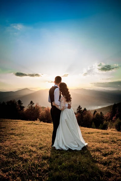 Noiva e noivo observam o pôr do sol em pé na colina — Fotografia de Stock