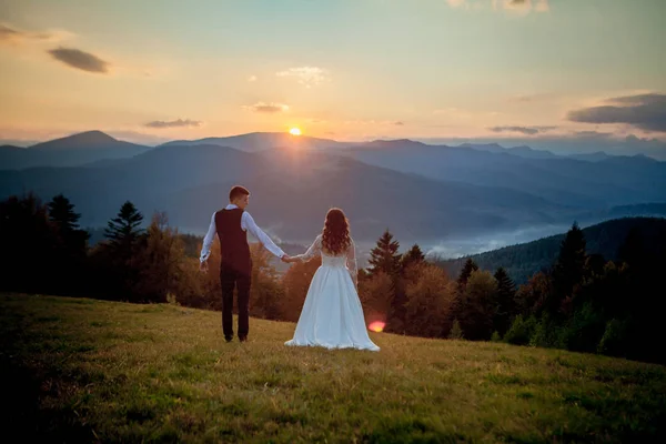 Noiva e noivo observam o pôr do sol em pé na colina — Fotografia de Stock