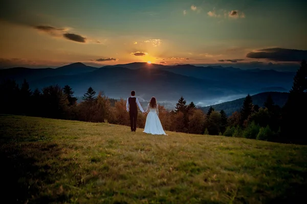 Noiva e noivo observam o pôr do sol em pé na colina — Fotografia de Stock