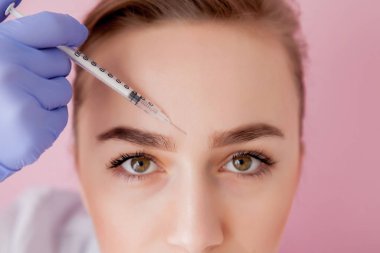 Doktor kozmetik uzmanı güzellik salonundaki genç ve güzel bir kadının cildindeki kırışıklıkları sıkmak ve düzleştirmek için yüz enjeksiyonlarını yenileme prosedürünü uyguluyor.