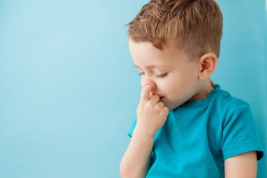 Küçük beyaz çocuk mavi arka planda burnunu karıştırıyor.