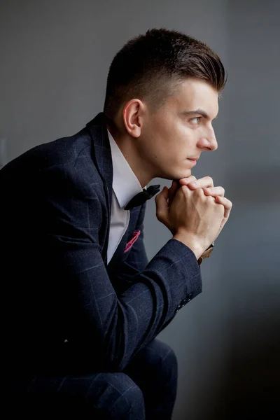 Concerned groom playing with his hands and looking to the side while wearing tuxedo, sitting on black studio background — Stock Photo, Image