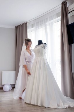 İpek bornozlu genç kadın gelinliğine dokunuyor ve pencerenin yanında dururken gülümsüyor. Düğün günü. Güzellik, moda. En mutlu düğün günü. Her anın tadını çıkar.