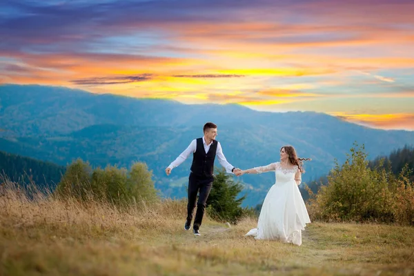 Boldog gyönyörű menyasszony és elegáns vőlegény futás és szórakozás, esküvő pár, luxus szertartás a hegyekben csodálatos kilátással — Stock Fotó
