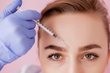 Doktor kozmetik uzmanı güzellik salonundaki genç ve güzel bir kadının cildindeki kırışıklıkları sıkmak ve düzleştirmek için yüz enjeksiyonlarını yenileme prosedürünü uyguluyor.