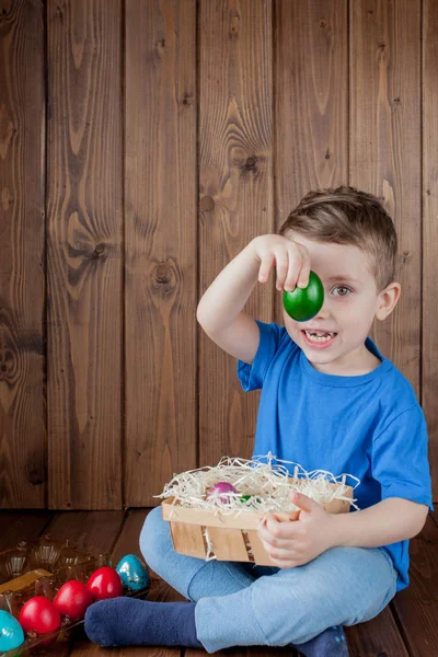 Happy baby boy with a basket of easter eggs on wooden background — Stock Photo, Image