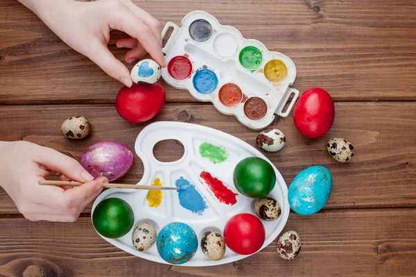 Hand painted Easter eggs, paints and brushes on wooden background. Preparation for the holiday. Girls hands draw a pattern