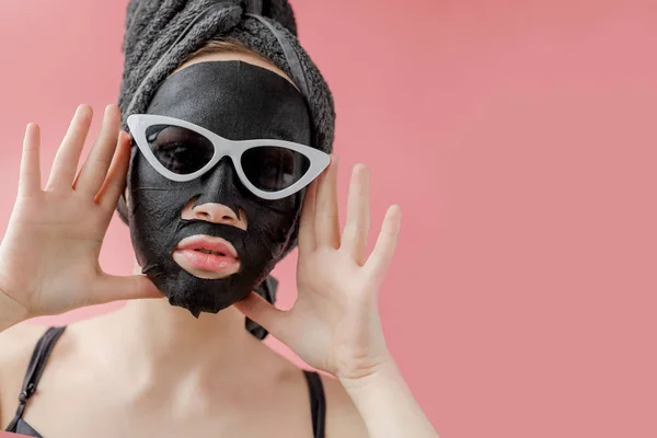 戴眼镜的年轻女子在粉色背景上涂上黑色化妆品织物的面膜 面部剥皮面膜用木炭 温泉美容治疗 靠近点 — 图库照片