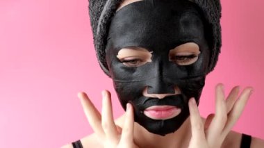 Genç bir kadın pembe arka plana siyah kozmetik yüz maskesi takıyor. Kömürlü maske soyma, kaplıca güzellik tedavisi, cilt bakımı, kozmetoloji. Kapat.