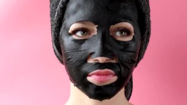 Genç bir kadın pembe arka plana siyah kozmetik yüz maskesi takıyor. Kömürlü maske soyma, kaplıca güzellik tedavisi, cilt bakımı, kozmetoloji. Kapat.