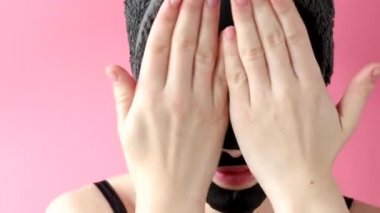 Genç bir kadın pembe arka plana siyah kozmetik yüz maskesi takıyor. Kömürlü maske soyma, kaplıca güzellik tedavisi, cilt bakımı, kozmetoloji. Kapat.