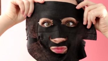 Genç bir kadın pembe arka plana siyah kozmetik yüz maskesi takıyor. Kömürlü maske soyma, kaplıca güzellik tedavisi, cilt bakımı, kozmetoloji. Kapat.