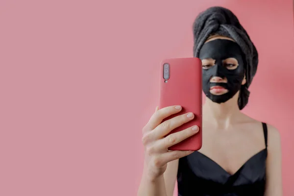Fiatal nő alkalmazni fekete kozmetikai szövet arc maszk és telefon a kezében rózsaszín háttér. Arctisztító maszk faszénnel, spa szépségápolás, bőrápolás, kozmetika. Közelről. — Stock Fotó