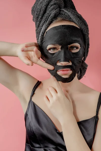 Mujer joven aplicar máscara facial de tela cosmética negro sobre fondo rosa. Mascarilla de descamación facial con carbón vegetal, tratamiento de belleza spa, cuidado de la piel, cosmetología. De cerca. — Foto de Stock