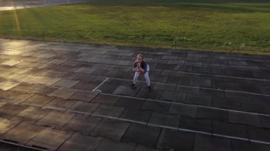 Spor giyimli genç zayıf bir kadın siyah kaplamalı bir stadyumda lastik bantla çömelme egzersizi yapıyor.