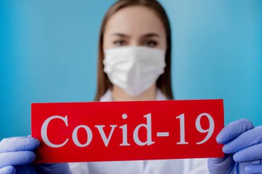 Cerrahi maskeli doktor kadın mavi arka planda kurtarıcı Coronavirüs olan kırmızı kağıda işaret ediyor. Dünya Sağlık Örgütü WHO, COVID-19 adlı Coronavirus hastalığına yeni bir resmi isim verdi.