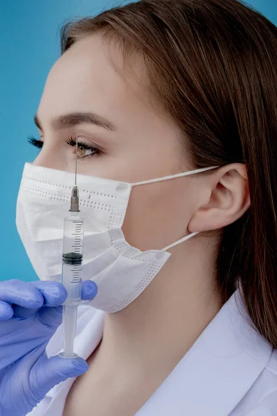 Woman doctor in a white medical coat, mask, hat and gloves holds a syringe with medicine in her hands. Standing on a white background.