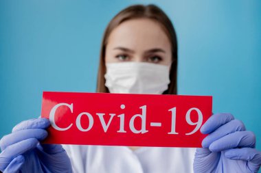 Cerrahi maskeli doktor kadın mavi arka planda kurtarıcı Coronavirüs olan kırmızı kağıda işaret ediyor. Dünya Sağlık Örgütü WHO, COVID-19 adlı Coronavirus hastalığına yeni bir resmi isim verdi.