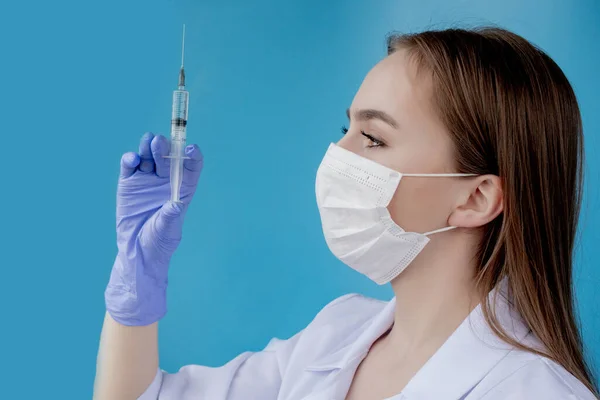 Woman doctor in a white medical coat, mask, hat and gloves holds a syringe with medicine in her hands. Standing on a white background.