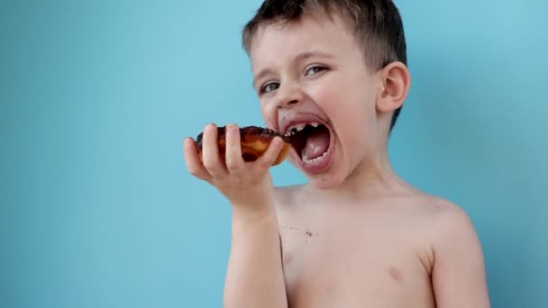 青い背景にドーナツチョコレートを食べる男の子 かわいい幸せな少年は口の周りにチョコレートで微笑んだ 子供のコンセプト 子供のためのおいしい食べ物 — ストック動画