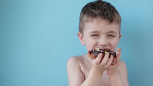 青い背景にドーナツチョコレートを食べる男の子 かわいい幸せな少年は口の周りにチョコレートで微笑んだ 子供のコンセプト 子供のためのおいしい食べ物 — ストック動画