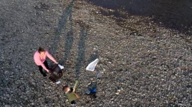 Oğluyla birlikte genç bir kadın nehirdeki bir çöp torbasında plastik çöp topluyor. Kullanılmış kirli plastik şişeler. Çevre kirliliği kıyısı