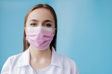 Doktor hemşire, cerrah maskesinin arkasında gülümsüyor. Mavi arka planda beyaz kadın modelin yakın plan portresi. Covid-19 'u korumak için kitle giyen kadın.