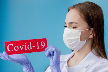 Cerrahi maskeli doktor kadın mavi arka planda kurtarıcı Coronavirüs olan kırmızı kağıda işaret ediyor. Dünya Sağlık Örgütü WHO, COVID-19 adlı Coronavirus hastalığına yeni bir resmi isim verdi.