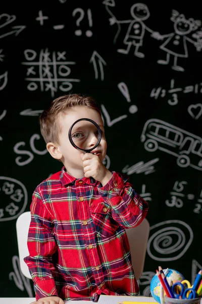 可愛い賢い少年は虫眼鏡を手に机に座っている。子供は背景に黒板が書かれた本を読んでいる。学校の準備だ。学校に戻る — ストック写真