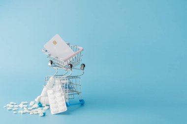 Dağılmış çeşitli haplar, ilaçlar, kısırlık, şişeler, termometre, şırınga ve mavi arka planda boş alışveriş arabası. Eczane alışveriş kavramı.