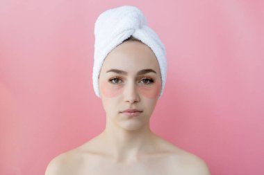 Kafasında beyaz havlu olan, göz altı kolajen bantlı pembe arka planda çıplak duran beyaz çilli kadının stüdyo fotoğrafı. Cilt bakımı, fotokopi alanı ile kozmetik ürün kavramı.