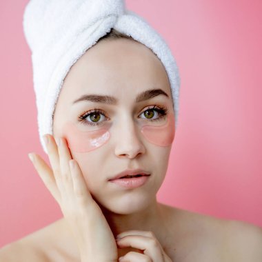 Kafasında beyaz havlu olan, göz altı kolajen bantlı pembe arka planda çıplak duran beyaz çilli kadının stüdyo fotoğrafı. Cilt bakımı, kozmetik ürün konsepti.