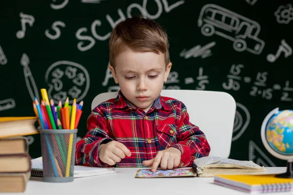 可愛い賢い少年は虫眼鏡を手に机に座っている 子供は背景に黒板が書かれた本を読んでいる 学校の準備だ 学校に戻る — ストック写真