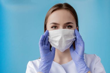 Doktor hemşire, cerrah maskesinin arkasında gülümsüyor. Mavi arka planda beyaz kadın modelin yakın plan portresi. Covid-19 'u korumak için kitle giyen kadın..
