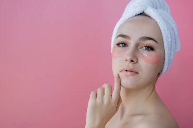 Kafasında beyaz havlu olan, göz altı kolajen bantlı pembe arka planda çıplak duran beyaz çilli kadının stüdyo fotoğrafı. Cilt bakımı, fotokopi alanı ile kozmetik ürün kavramı.