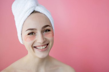 Kafasında beyaz havlu olan, göz altı kolajen bantlı pembe arka planda çıplak duran beyaz çilli kadının stüdyo fotoğrafı. Cilt bakımı, fotokopi alanı ile kozmetik ürün kavramı.