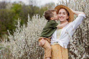 Mutlu anne ve oğul birlikte eğleniyor. Anne oğluna nazikçe sarılıyor. Arka planda beyaz çiçekler açar. Anneler Günü..