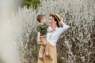 Mutlu anne ve oğul birlikte eğleniyor. Anne oğluna nazikçe sarılıyor. Arka planda beyaz çiçekler açar. Anneler Günü..