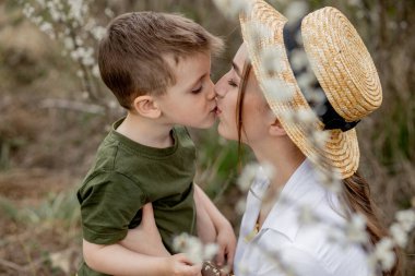 Mutlu anne ve oğul birlikte eğleniyor. Anne oğluna nazikçe sarılıyor. Arka planda beyaz çiçekler açar. Anneler Günü..